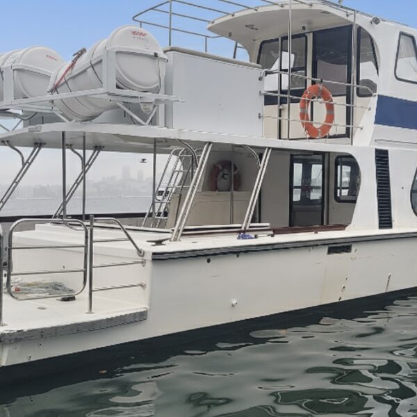 A white motorboat with two decks and several life rings floats on calm water, with a city skyline visible in the distance under a blue sky.