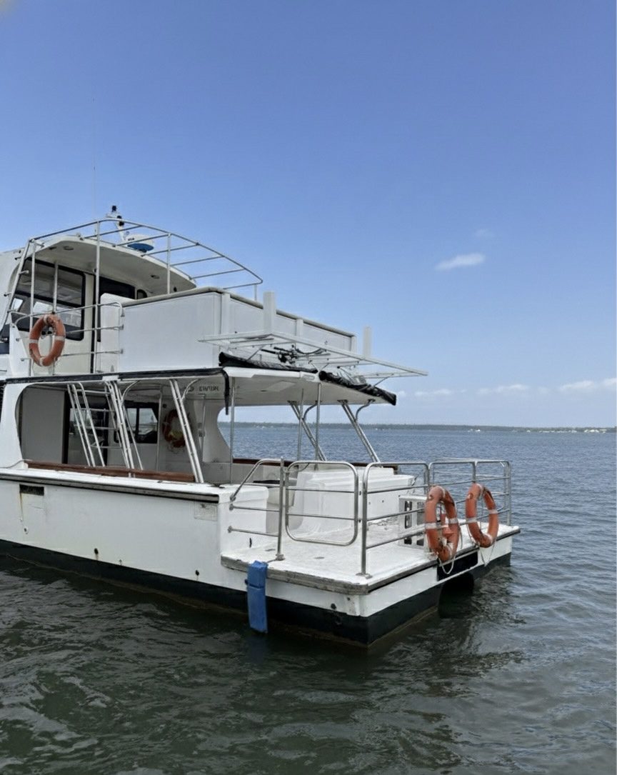 A white motorboat with orange lifebuoys is docked on calm water under a clear blue sky. The boat has two decks and metal railings, with distant land visible on the horizon.