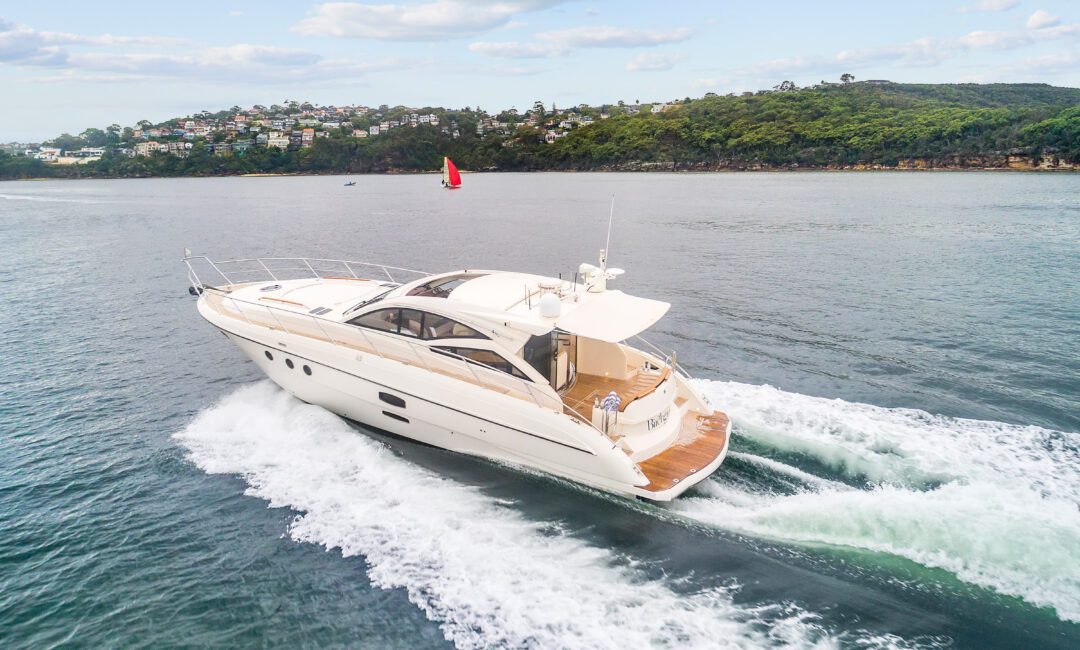 Birchgrove, a white luxury yacht, speeds through calm waters near a green, hilly coast under partly cloudy skies, leaving a wake behind; houses dot the distant hillside.
