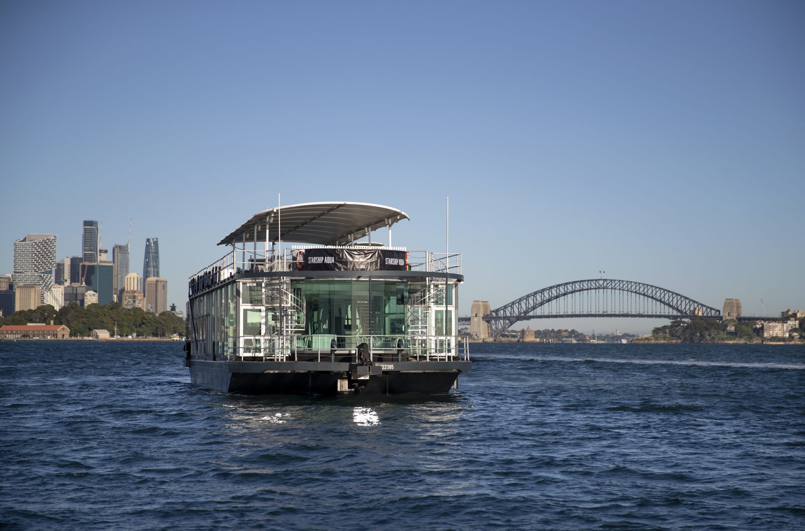 A modern glass-walled boat floats on blue water, with Sydney’s skyline and the Sydney Harbour Bridge visible in the background under a clear blue sky.