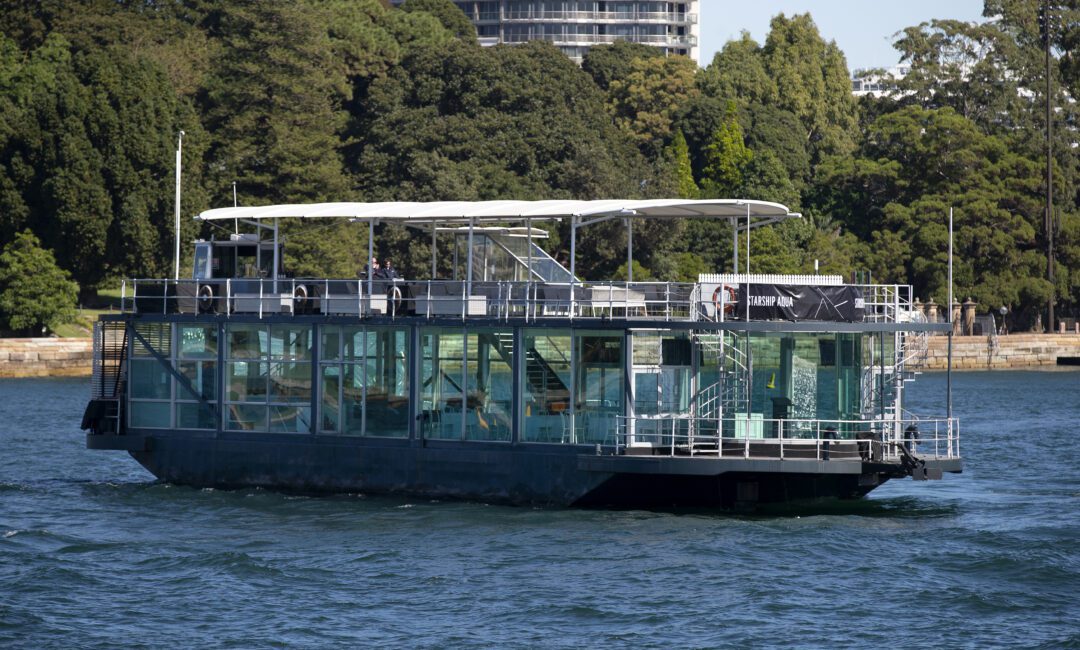 A modern glass-walled boat with a white canopy sails on a body of water, with a lush green park and tall buildings visible in the background.