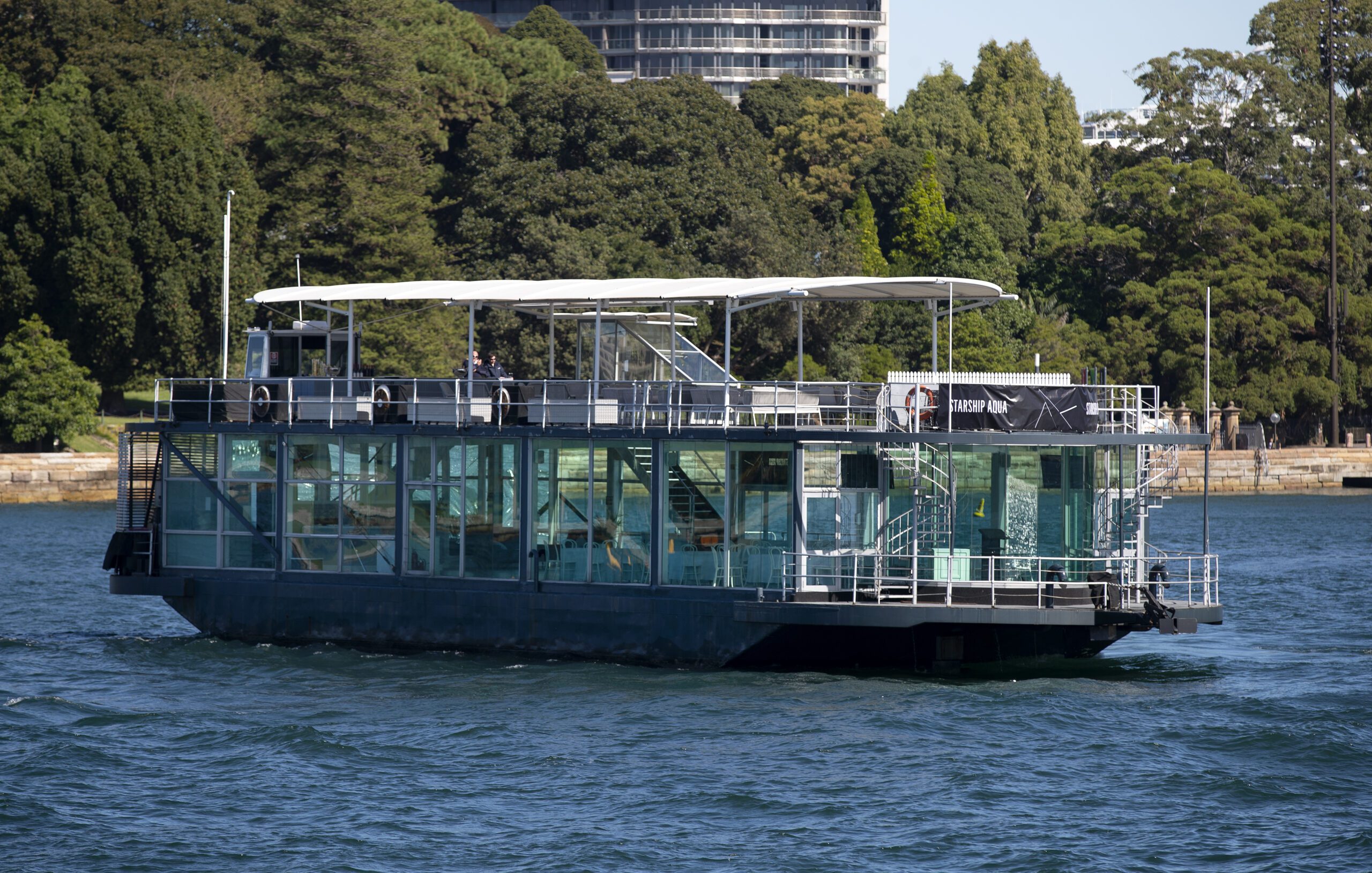 A modern glass-walled boat with a white canopy sails on a body of water, with a lush green park and tall buildings visible in the background.