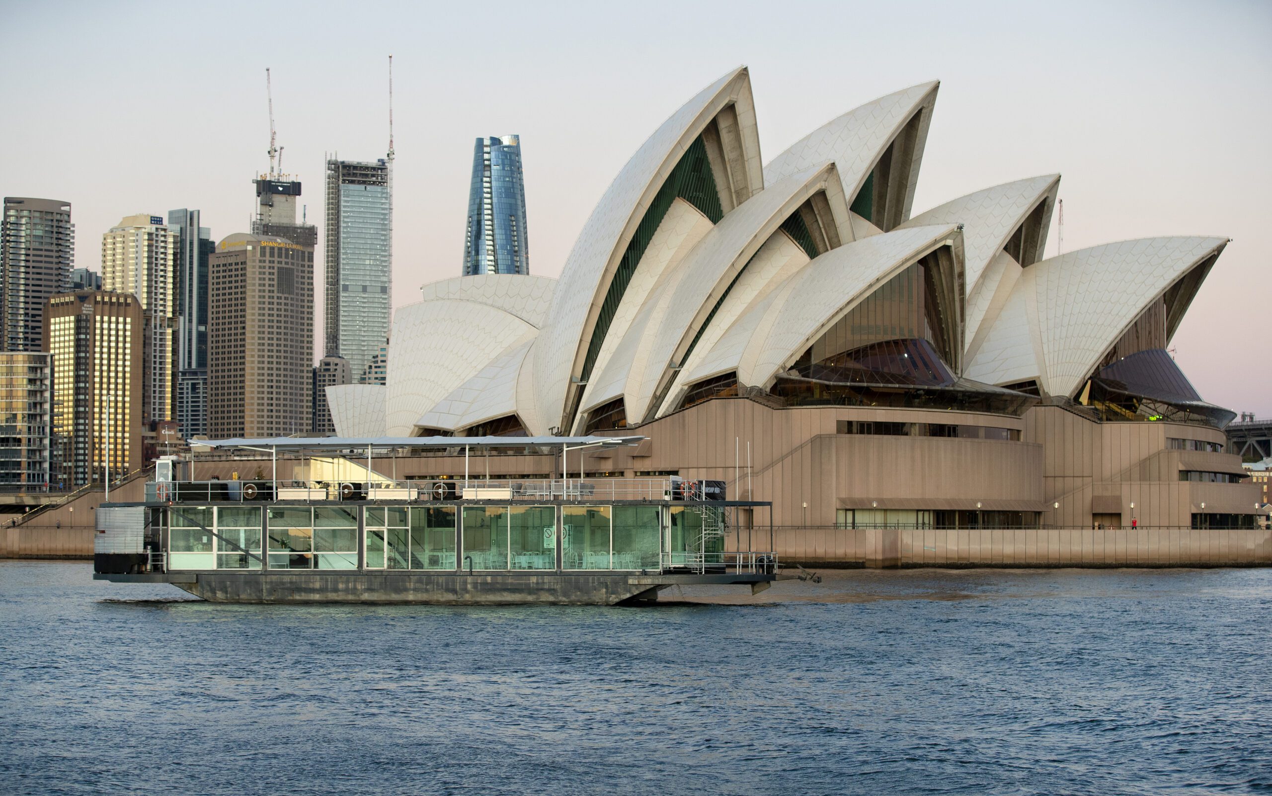 A modern glass-walled boat floats on the water in front of the iconic Sydney Opera House, with city skyscrapers visible in the background at dusk.