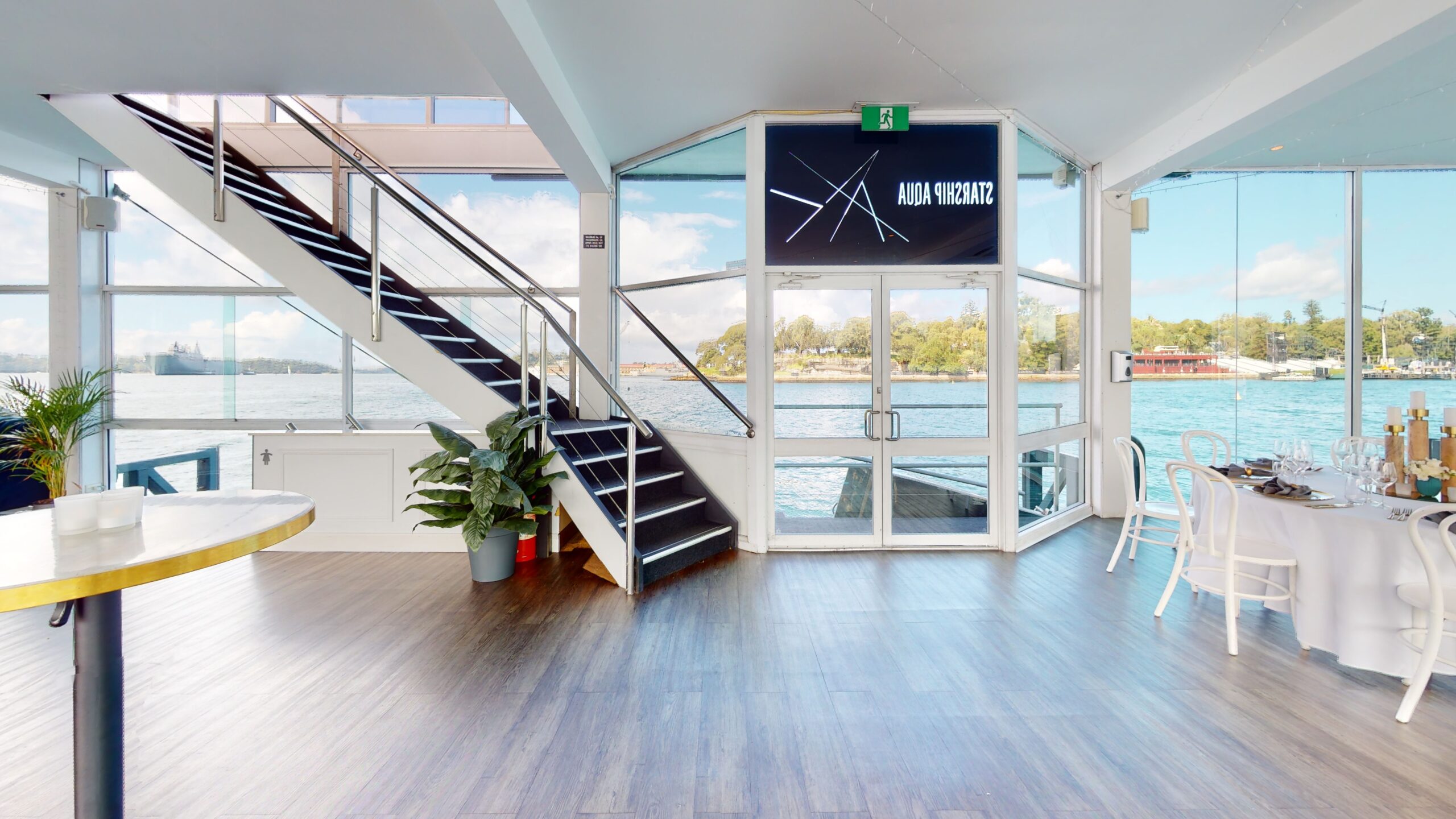 A bright, modern interior with large glass doors and windows overlooking the water, a black metal staircase, indoor plants, and a table set for dining near the right side. A sign above the door reads "AALIA VAGANZA.