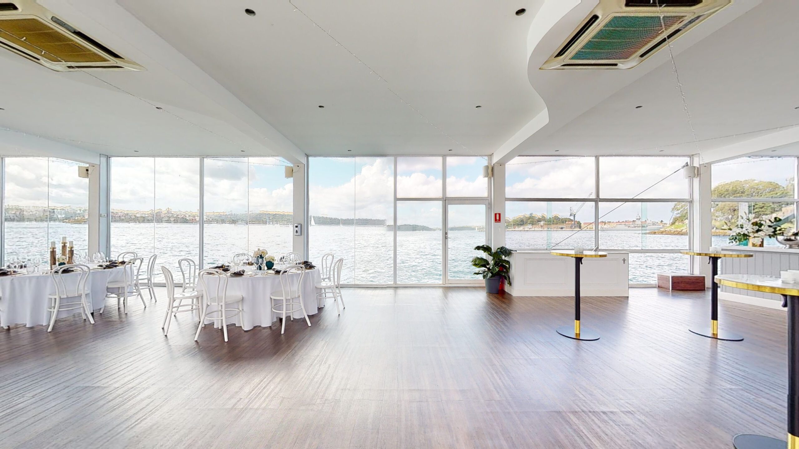 A bright, spacious event hall with large floor-to-ceiling windows overlooking a body of water, featuring round tables set for dining on the left and tall cocktail tables on the right.