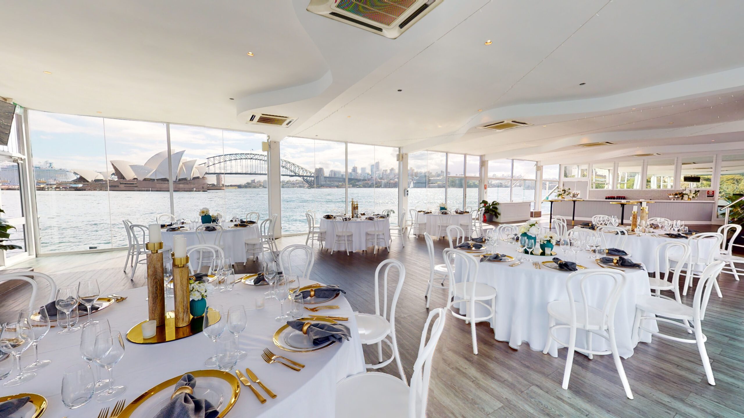 Elegant waterfront banquet hall with round tables set for an event, gold and teal decor, large windows showing views of Sydney Opera House and Harbour Bridge across the water.
