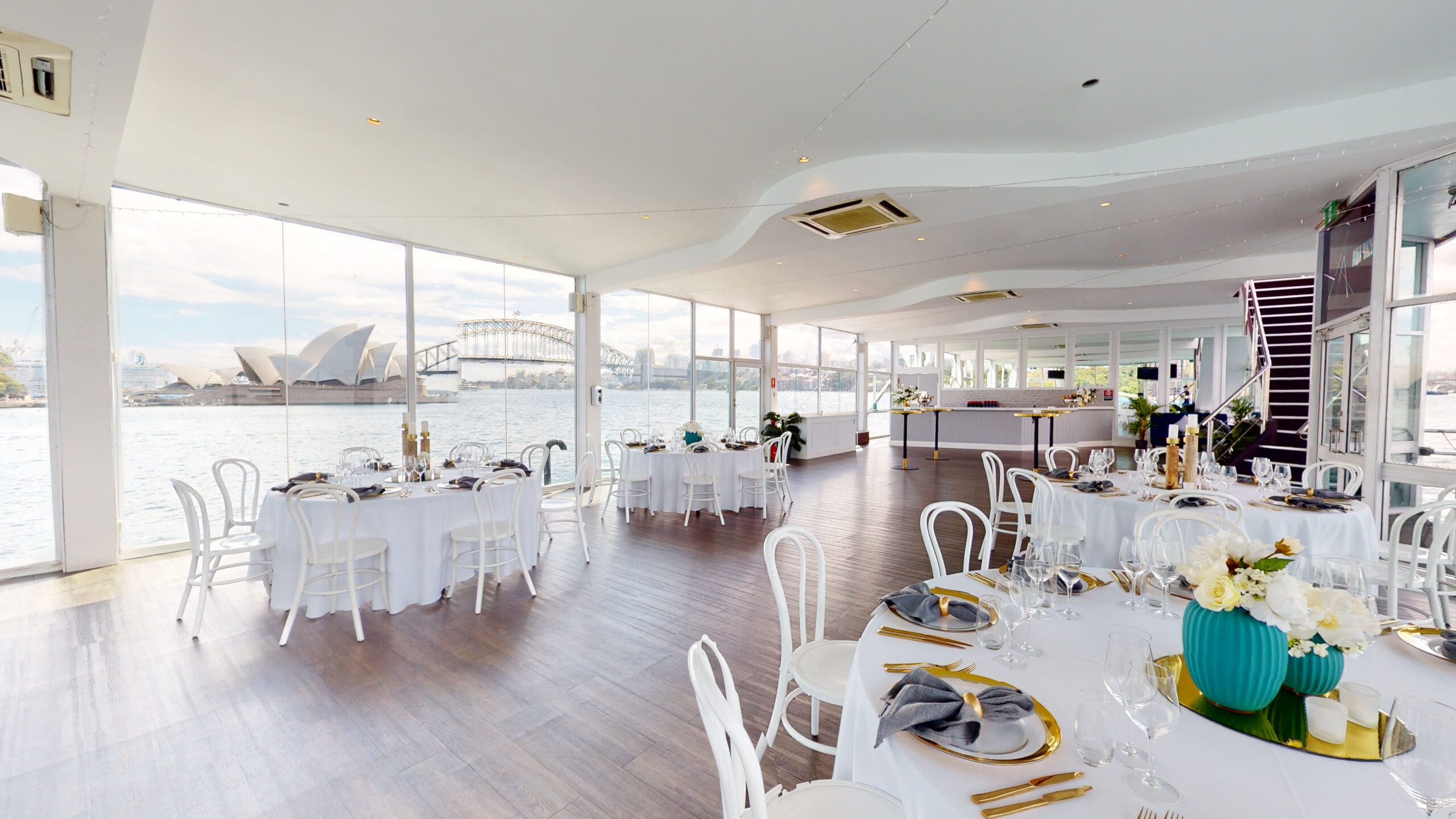 A bright, modern event space with round tables set for dining, white chairs, large windows overlooking the Sydney Opera House and Harbour Bridge, and flowers as centerpieces.