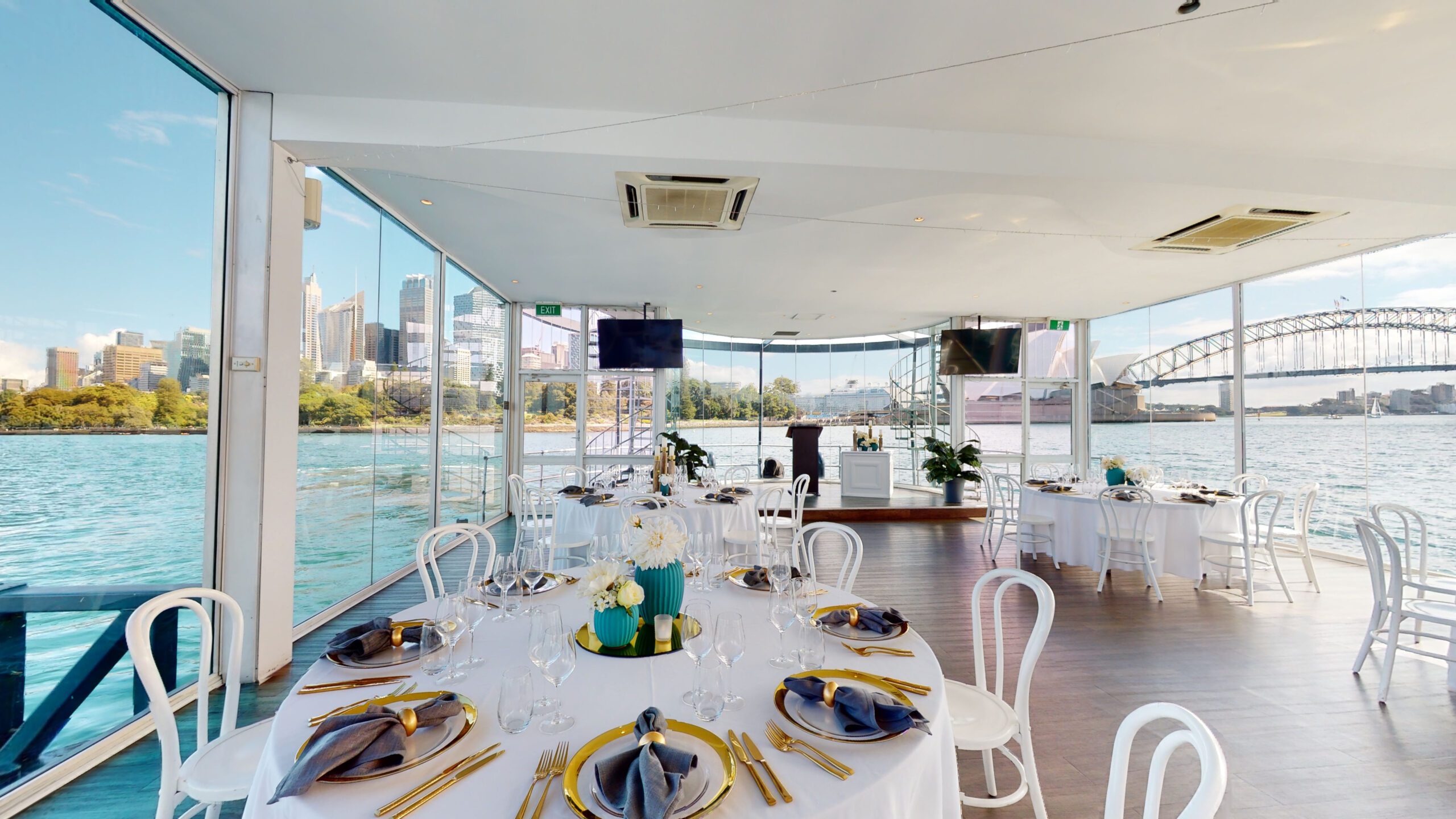 A bright, modern dining area on a boat with large windows showing city buildings and a bridge over water. Tables are elegantly set with gold cutlery, white chairs, and floral centerpieces.