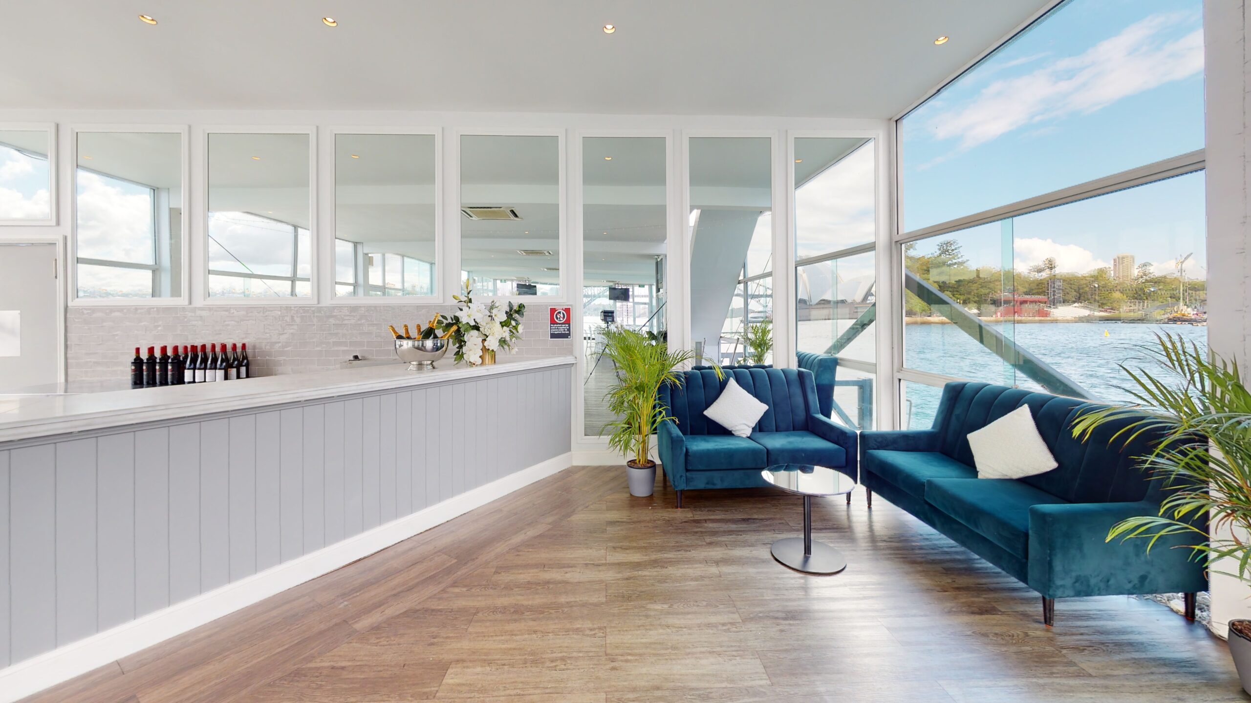 A modern lounge with blue velvet sofas, potted plants, and a round glass table beside large windows overlooking water. A sleek bar with wine bottles and flowers is on the left, with wood flooring throughout.
