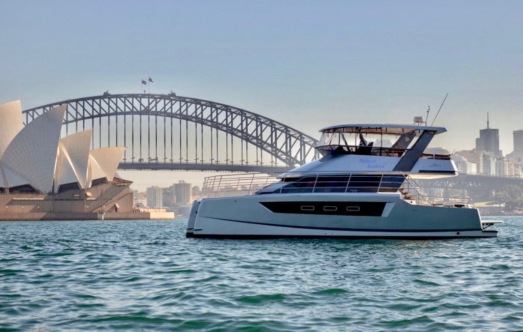 A modern yacht floats on the water with the Sydney Opera House and Sydney Harbour Bridge in the background under a clear sky.