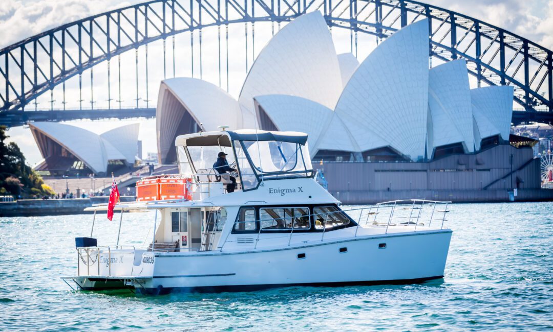 A white yacht named "Enigma X" sails on Sydney Harbour, with the Sydney Opera House and Harbour Bridge visible in the background under a partly cloudy sky.