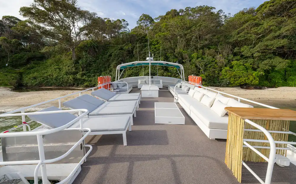 The Carribean Cat’s outdoor upper deck features white lounge chairs, sofas, a small table, and a bamboo bar, all overlooking a sandy beach with lush green trees in the background.