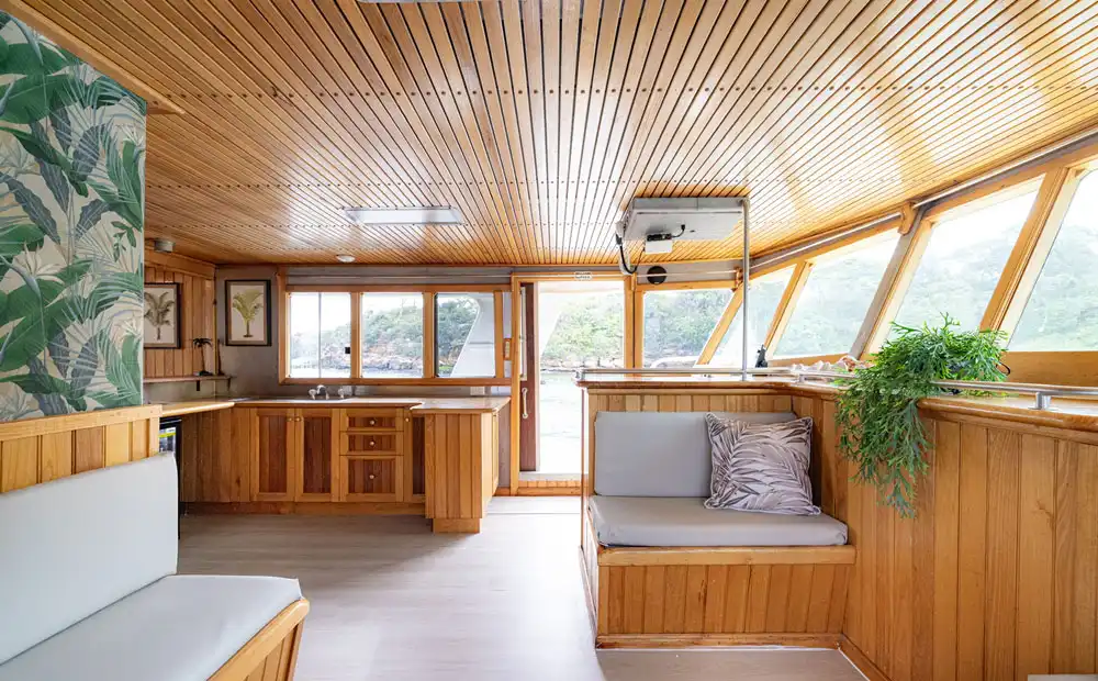 Bright, modern houseboat interior with wood paneling, large windows, cushioned built-in benches, potted plants, and a compact kitchen. Outside, lush greenery is perfect for relaxing with your Carribean Cat on sunny days.