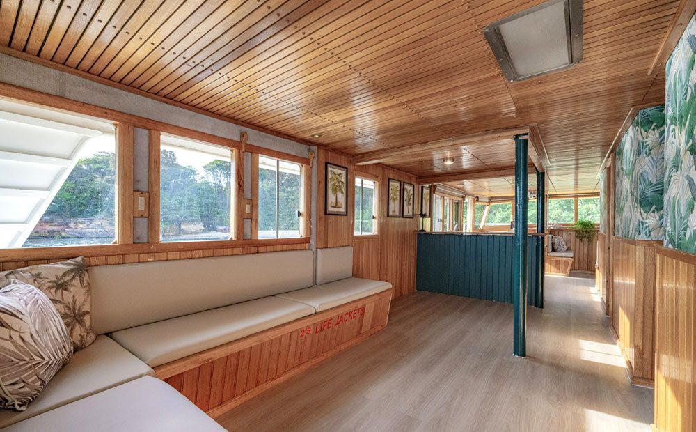 The Carribean Cat’s interior features wood-paneled walls and ceiling, large windows, cushioned seating with decorative pillows, framed art, and views of trees and water, filling the spacious area with natural light and a welcoming ambiance.