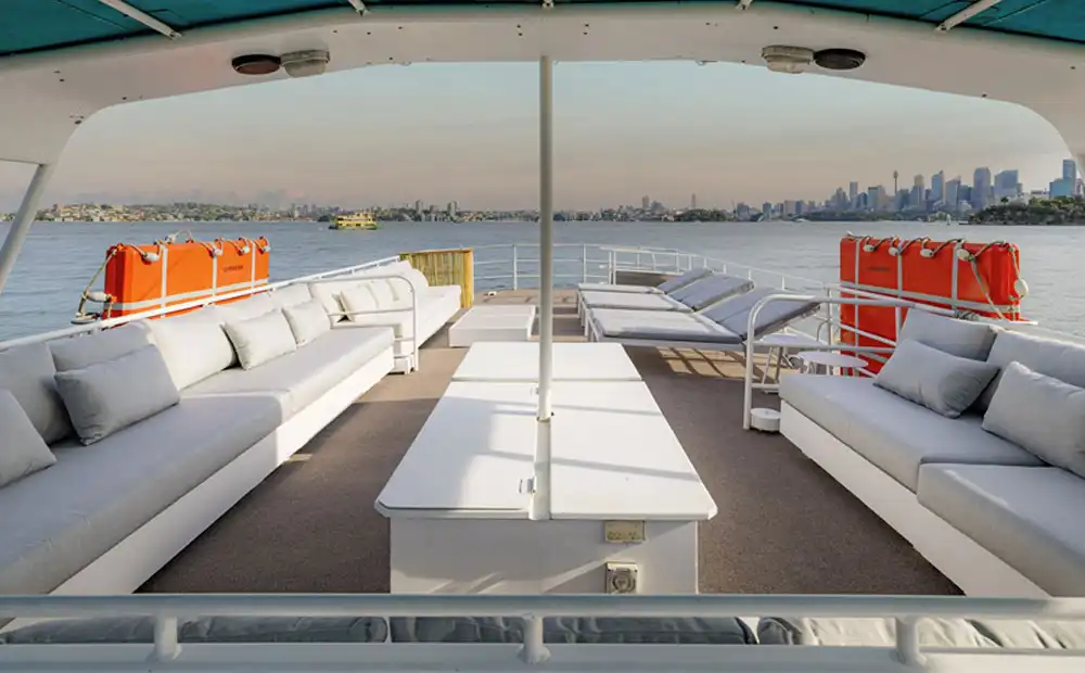 The Carribean Cat features a spacious deck with white cushioned seating, tables, and orange life preservers on both sides, offering views of calm water and a distant city skyline under a clear sky.