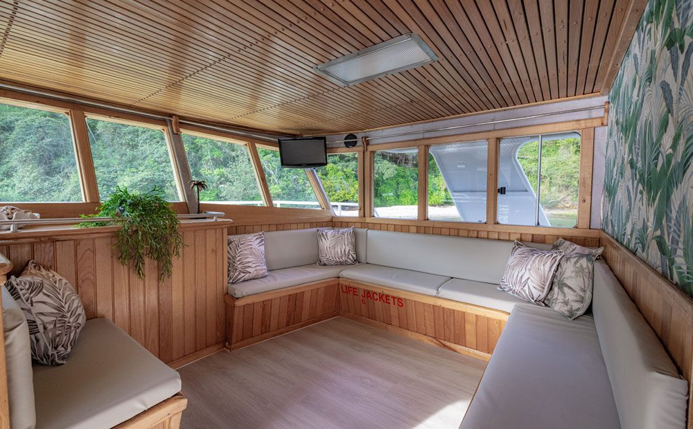 The Carribean Cat features a cozy interior with light wood paneling, bench seating with patterned cushions, large windows showcasing greenery, a small TV, a counter plant, and "LIFE JACKETS" labeled in red under a seat.