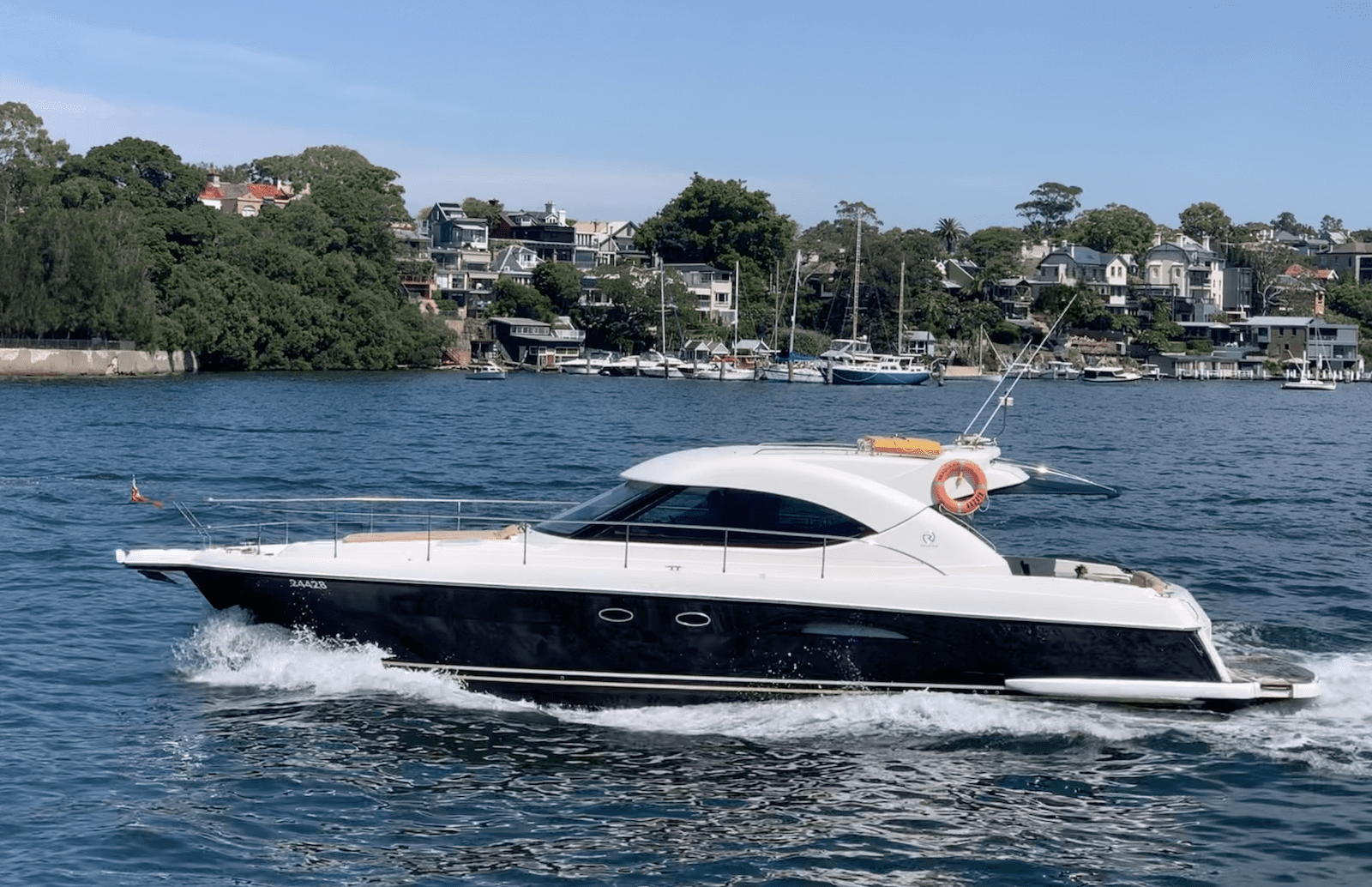 A sleek black and white motor yacht cruises on calm blue water, with a row of houses, trees, and moored boats visible along the shoreline in the background under a clear sky.