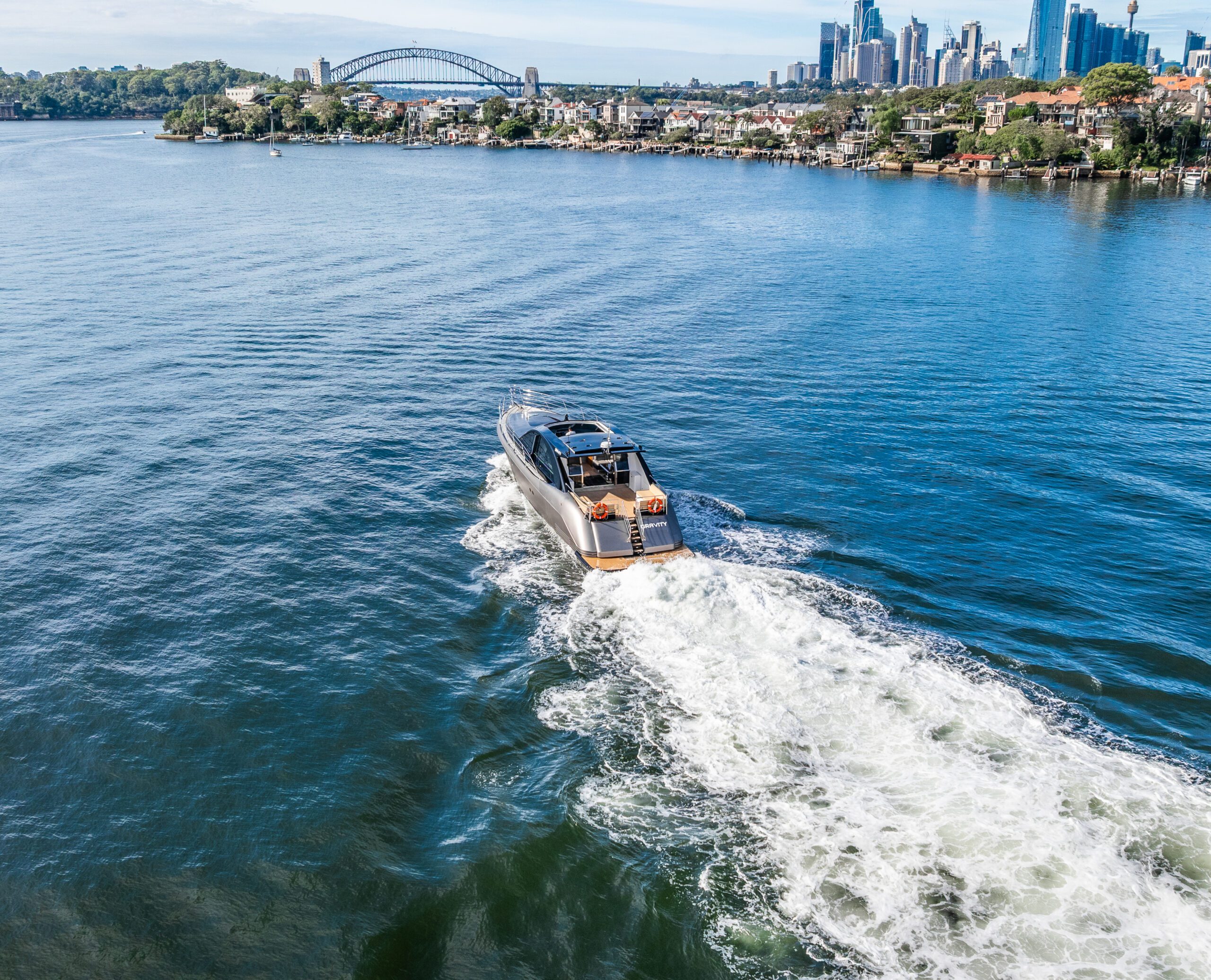 A motorboat creates a white wake as it speeds across a wide river, with the Sydney Harbour Bridge and city skyline visible in the background under a clear blue sky.