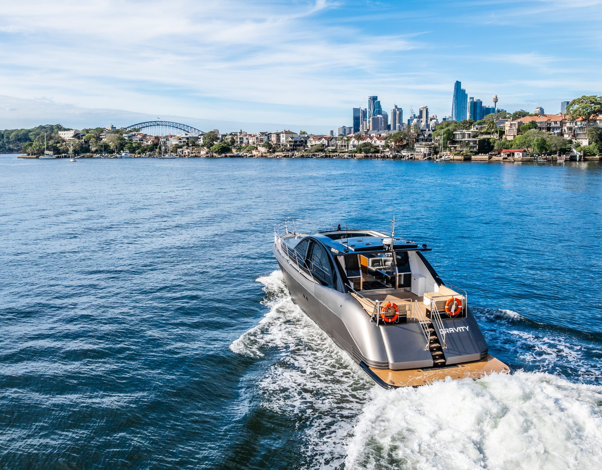 A sleek modern yacht cruises on blue water toward a city skyline under a partly cloudy sky, with a bridge and waterfront houses visible in the background.