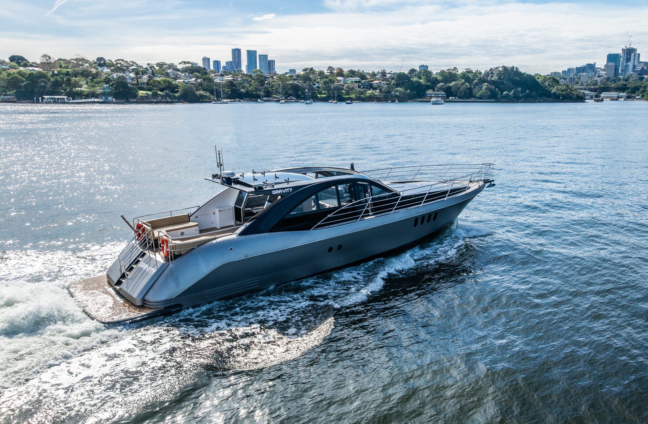 A sleek, modern yacht cruises through calm blue water near a city shoreline, with trees and tall buildings visible in the background under a partly cloudy sky.