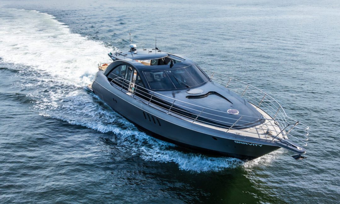 A sleek, modern motor yacht named "GRAVITY" cruises across calm blue water near a shoreline with trees and houses in the background.