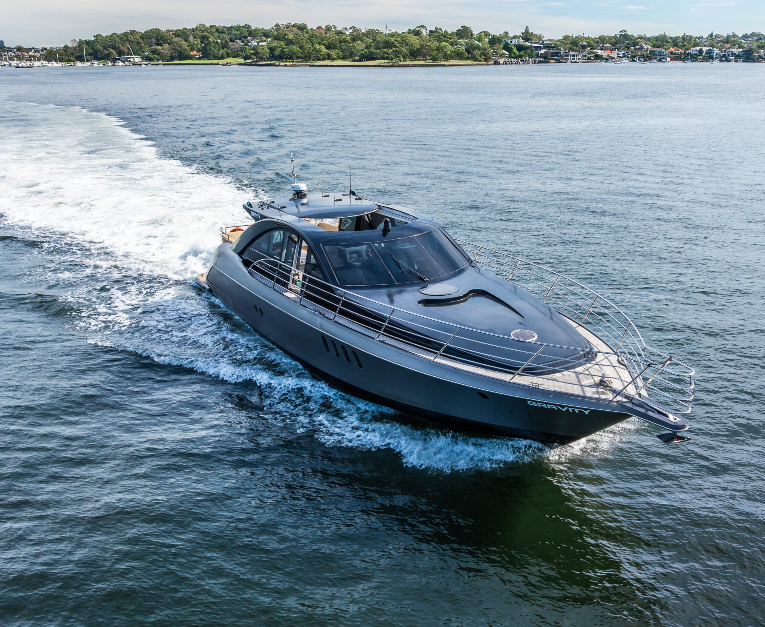 A sleek, modern motor yacht named "GRAVITY" cruises across calm blue water near a shoreline with trees and houses in the background.