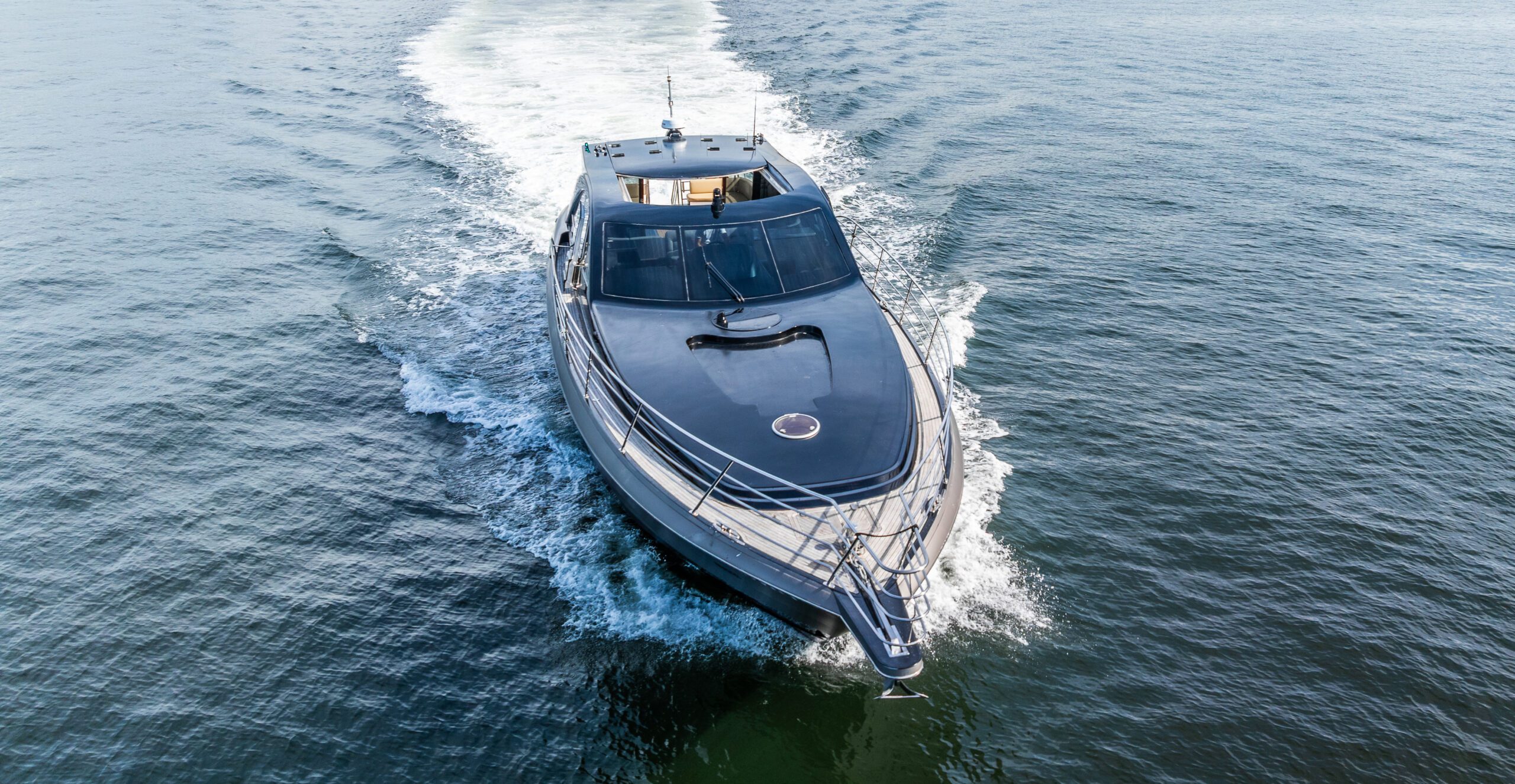 A sleek, modern yacht moves swiftly across calm blue water, creating a white wake behind it under a clear sky. The photo is taken from above, showing the yacht’s streamlined design and spacious deck.