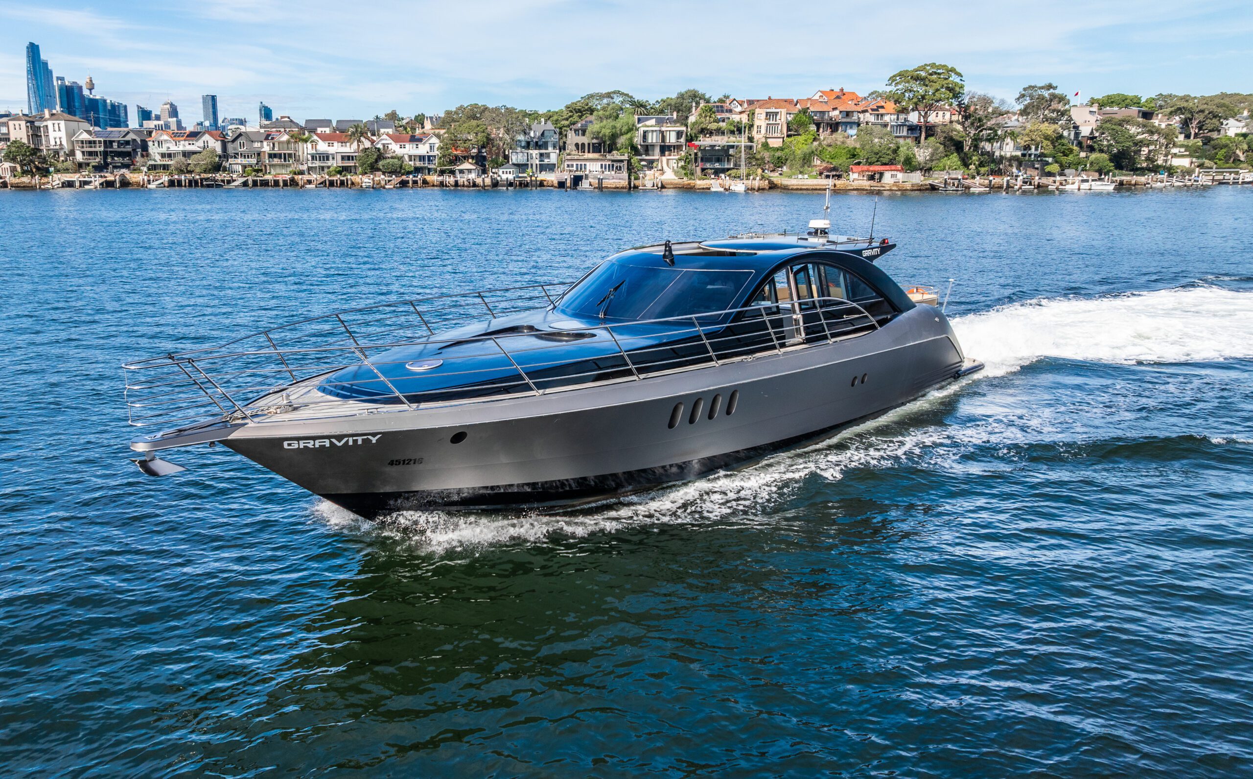 A sleek, modern black yacht named "GRAVITY" cruises on calm blue water, leaving a trail of white waves, with a shoreline of houses and trees in the background under a clear sky.