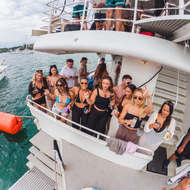 A group of people in swimwear enjoy drinks and music on the deck of a boat, smiling and posing for a photo. The boat is on the water with other boats and a cloudy sky in the background.