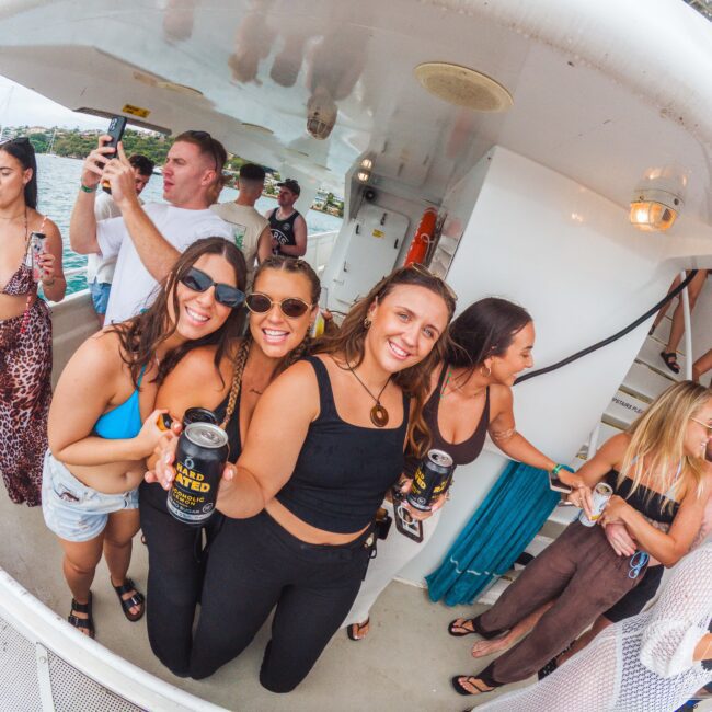 A group of young adults smiling and holding drinks on a boat. Some are taking photos, while others chat. They're dressed in summer outfits, and the water and other boats are visible in the background.