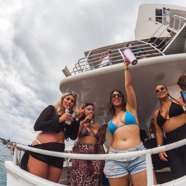 Four women in swimsuits stand on the deck of a boat, smiling and posing for the camera. One woman raises a drink in the air. The sky is cloudy, and the sea is visible in the background.