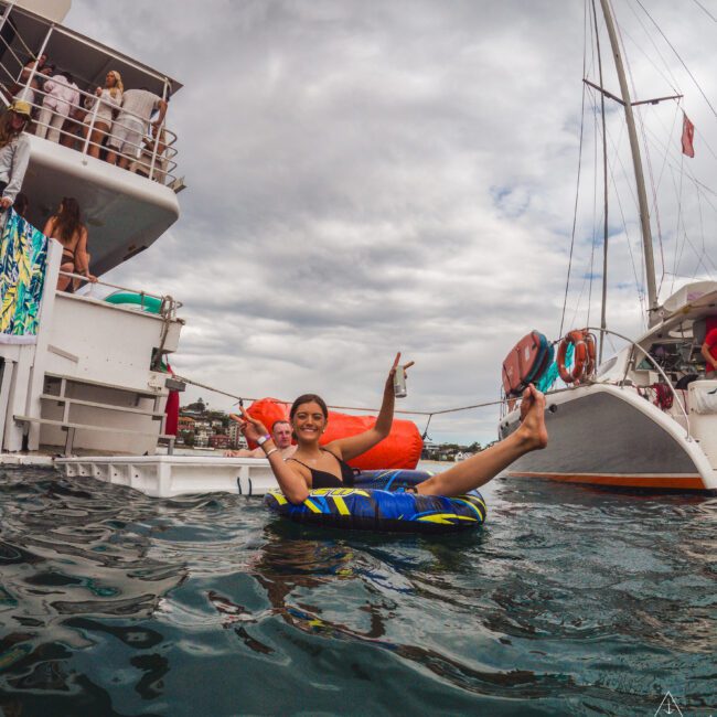 A smiling woman lounging in a blue inflatable tube floats between boats on the water, making a peace sign with her hand. Several people stand on a nearby boat under a cloudy sky.