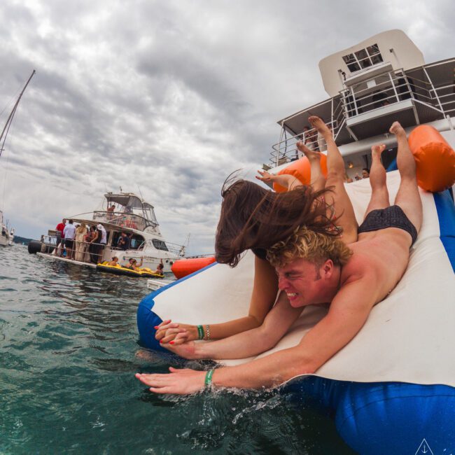 Two people slide down an inflatable slide from a yacht into the water, laughing and holding hands, with other boats and people visible in the background under a cloudy sky.