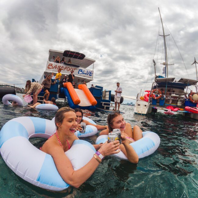 Three women in striped pool floats enjoy drinks in the water near boats. Other people relax on a floating platform labeled “Barefoot Blue” under a cloudy sky. The scene is festive and social.