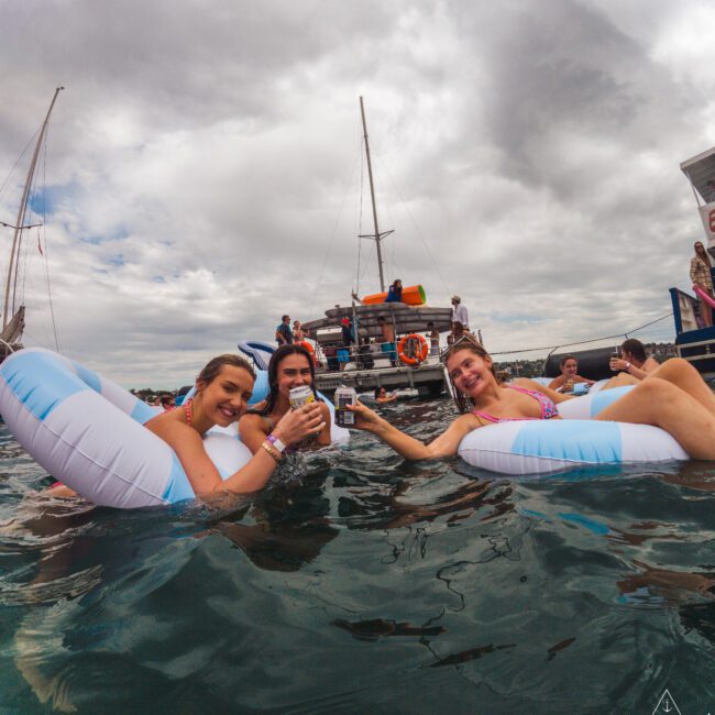 Three women relax on inflatable pool floats in the water, smiling and holding drinks, with boats and people in the background under a cloudy sky.