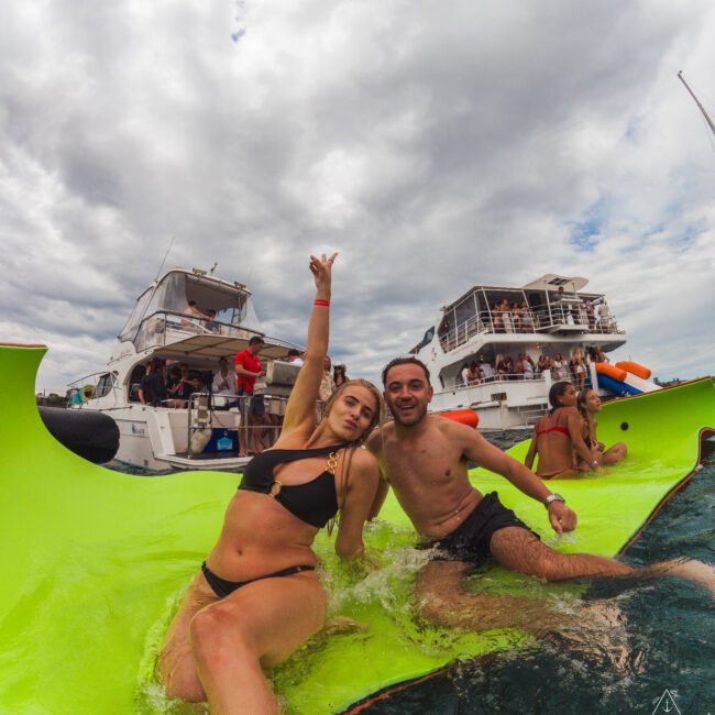 Two people in swimwear smile and pose on a bright green floating mat in the water, with boats and other people in the background under a cloudy sky.