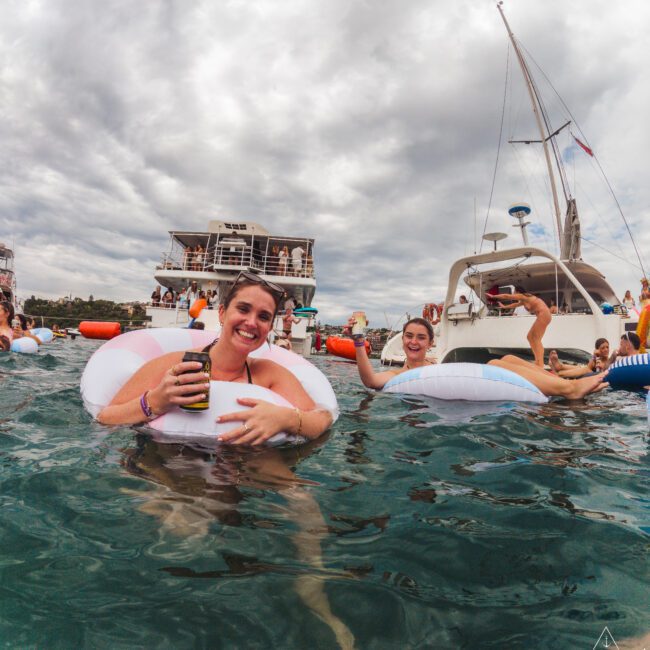 Two smiling women float in the water on inflatables, holding drinks, near several anchored boats with people partying onboard under a cloudy sky.