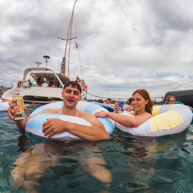 A man and woman relax on inflatable pool rings in the ocean, holding drinks, with a sailboat and other people in the background under a cloudy sky.