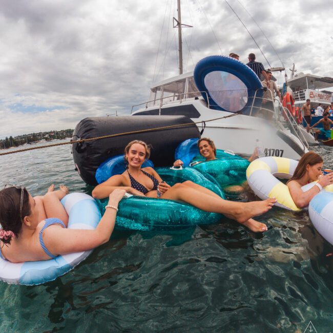 Four women relax on colorful inflatable floats in the water near a yacht, smiling and enjoying a sunny day. Other people and the boat are visible in the background under a partly cloudy sky.