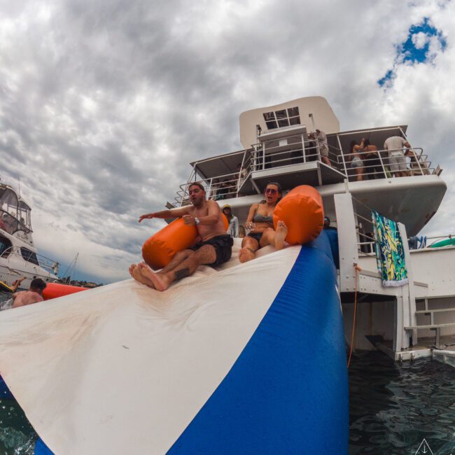 Two people in swimsuits slide down a large inflatable water slide attached to a boat, with others watching from the boat deck. The sky is cloudy and the scene appears lively and fun.