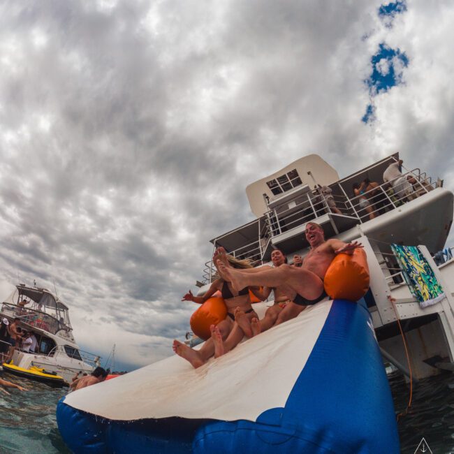 A group of people joyfully slide down an inflatable slide attached to a yacht, with other boats and cloudy skies visible in the background. The scene is lively and playful on the water.