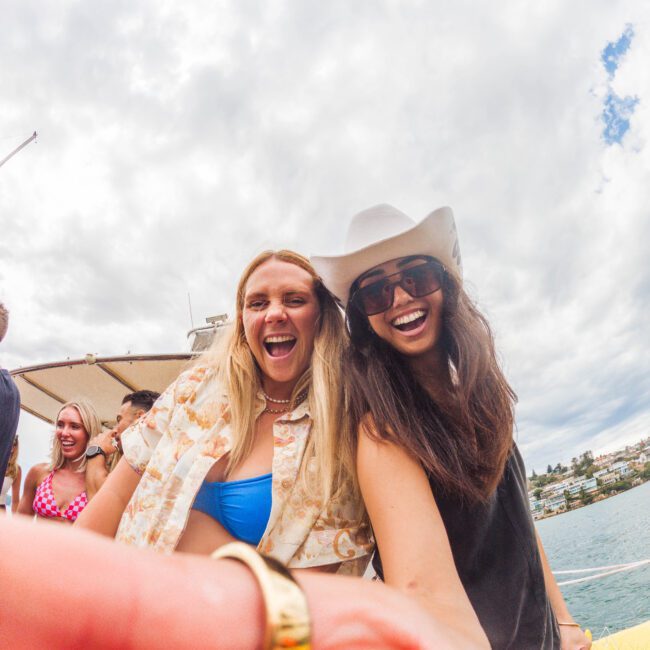Two smiling women pose for a selfie on a boat, one wearing a white hat and sunglasses, the other in a patterned shirt and blue bikini top. Other people and water are visible in the background under a cloudy sky.