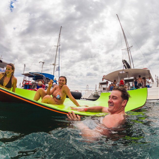 A man in the water smiles while holding onto a large floating mat with two women lounging on it. Sailboats and people are visible in the background under a cloudy sky.