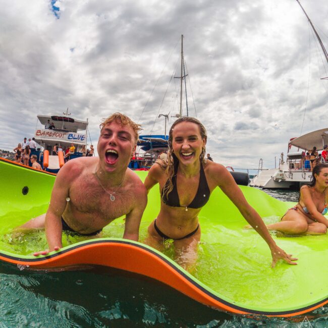 Two smiling young adults in swimsuits play on a floating mat in the ocean, surrounded by boats and people. The sky is cloudy, and both are laughing and enjoying the water.