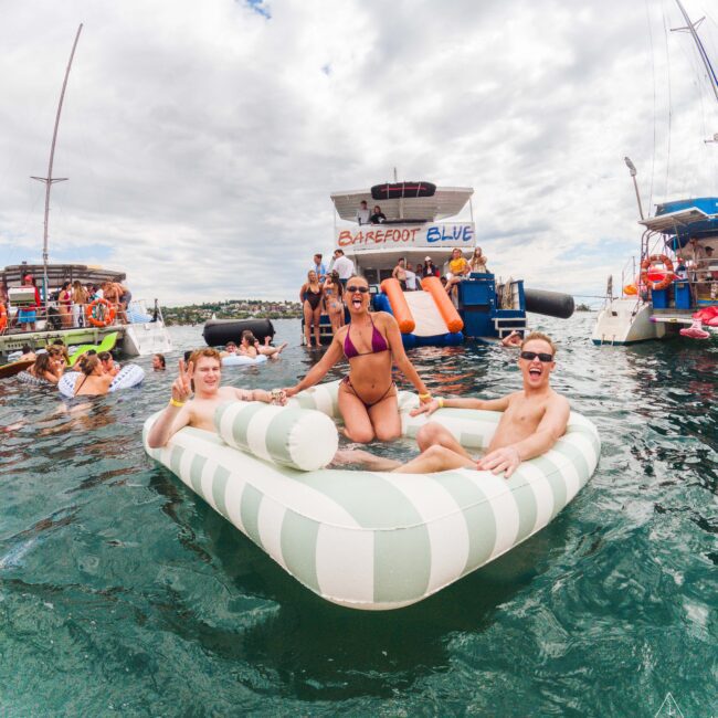 Three people smile and pose on a striped inflatable float in the water near several boats. Other people swim and socialize around them under a partly cloudy sky. The scene looks lively and festive.