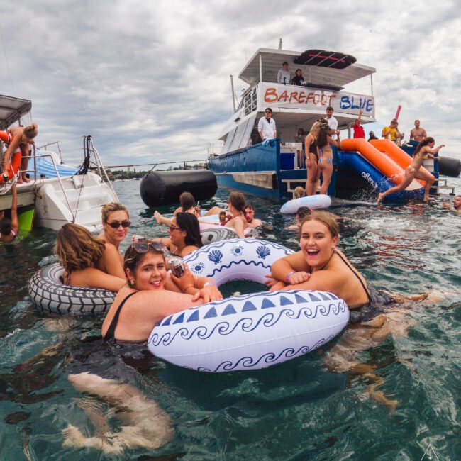 A group of people relax and socialize on colorful inflatable rings in the water near a boat named "Barefoot Blue" under a cloudy sky. Some are laughing and enjoying the lively atmosphere.