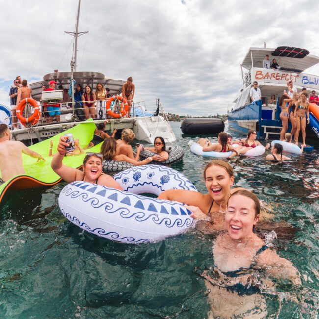 A group of smiling young adults relax in the water on pool floats near boats, with more people partying on deck under a cloudy sky. The scene is lively and festive.