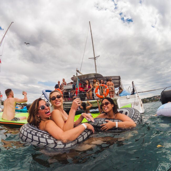 Three women in swimsuits smile and pose in an inflatable tube on the water near a boat with a group of people. The sky is cloudy, and the scene looks festive and cheerful.