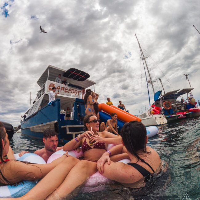 People relaxing on inflatable tubes in the water near a boat labeled "BAREFOOT," with others socializing on the deck. The sky is cloudy and several boats are anchored nearby. A bird is flying overhead.