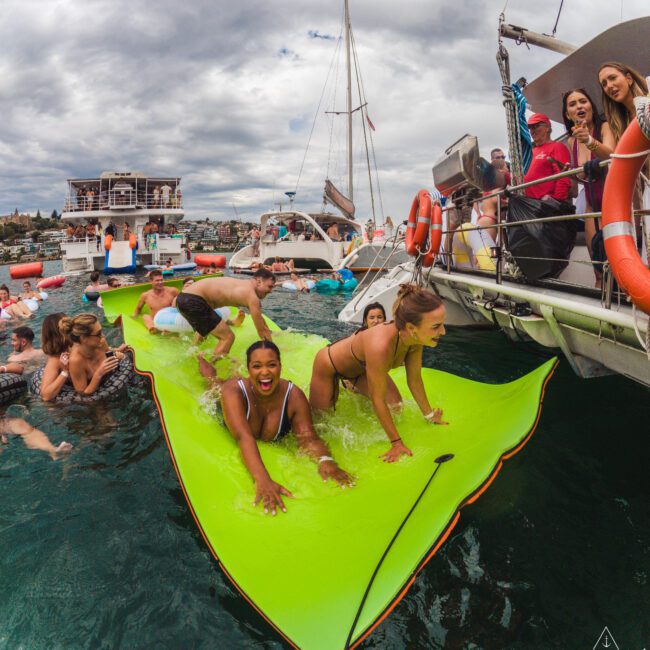 People are having fun on a bright green floating mat in the water next to a boat, laughing and playing as others watch and take photos, with more boats and a cloudy sky in the background.